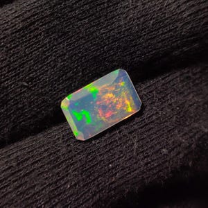 Könnte beinhalten: Ein rechteckiger, irisierender Opal-Edelstein mit einem grün-rot-gelben Farbspiel. Der Opal liegt auf einem schwarzen Stoffhintergrund.