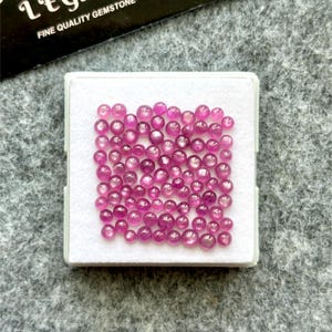 May include: A collection of small, round, pink gemstones displayed in a white tray. The gemstones are of varying sizes and have a polished, reflective surface. The tray is set against a gray background, with a partially visible black card.