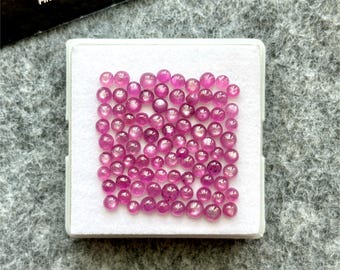 Pode incluir: Uma coleção de pequenas pedras preciosas redondas e rosa exibidas numa bandeja branca. As pedras preciosas, de vários tamanhos, têm uma superfície polida. A bandeja está sobre um fundo cinzento, com um cartão preto parcialmente visível.