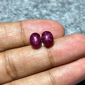 May include: Two oval, deep red gemstones with a star-like effect. The gems are held in a hand, showing their polished surfaces and rich colour. These gemstones are likely rubies, suitable for jewellery.