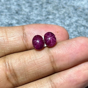 Könnte beinhalten: Zwei ovale, tiefrote Edelsteine mit sternförmigem Muster. Die Edelsteine liegen auf einer weißen Oberfläche, vor einem unscharfen Hintergrund. Die Edelsteine haben eine satte, dunkelrote Farbe.