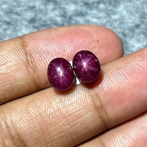 May include: A pair of oval-shaped cabochon star rubies with a deep magenta-red hue. Each gemstone displays a distinct six-rayed star effect, known as asterism, visible on the smooth, polished, and rounded surface of the stones.