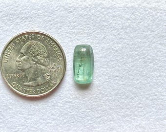 May include: A small, rectangular, translucent light green gemstone with rounded corners, positioned next to a United States quarter dollar coin for scale, showing the relative size of the stone compared to the coin's 0.955 inch diameter.