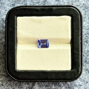 May include: A purple gemstone with a rectangular shape is displayed in a black velvet ring box.