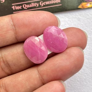 May include: Two oval-shaped, faceted pink gemstones with a translucent appearance. The stones feature a geometric cut with multiple triangular facets that reflect light, showcasing a vibrant magenta-pink hue throughout the polished surfaces.