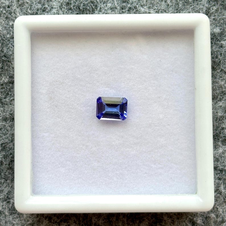 May include: A blue gemstone with a rectangular emerald cut, displayed in a white square box.