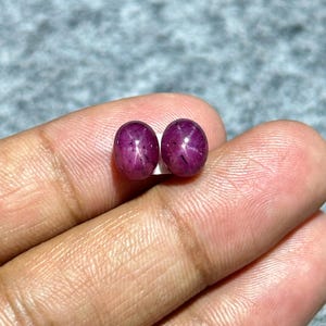 May include: Two oval-shaped, deep red star ruby gemstones. The gems have a polished surface with a visible star effect. The rubies are displayed on a light background.