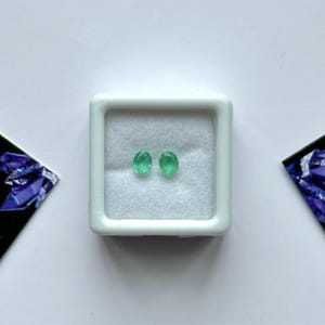 Puede incluir: Dos esmeraldas ovaladas de color verde brillante presentadas en una caja de joyer&iacute;a cuadrada blanca. Las piedras preciosas est&aacute;n colocadas sobre un fondo blanco. Tarjetas negras con un dise&ntilde;o geom&eacute;trico a cada lado de la caja.