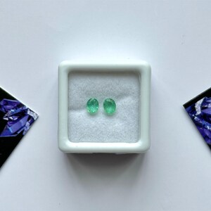 Puede incluir: Dos piedras preciosas de esmeralda ovaladas, de color verde brillante, se exhiben en una peque&ntilde;a caja de joyer&iacute;a cuadrada blanca. Las piedras preciosas est&aacute;n sobre un fondo blanco. La caja est&aacute; centrada en una superficie blanca, con tarjetas oscuras y anguladas con un dise&ntilde;o de cristal p&uacute;rpura a cada lado.