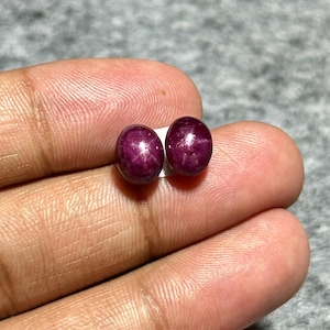 Może przedstawiać: Dwa owalne, głęboko czerwone kamienie szlachetne z efektem gwiazdy. Kamienie są prezentowane na białej powierzchni. Kamienie mają bogaty, ciemnoczerwony kolor i wydają się wypolerowane. Tło jest neutralnie szare.