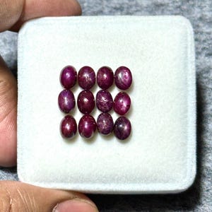 May include: A collection of 12 oval-shaped ruby gemstones, each with a star-like pattern, displayed in a white velvet-lined box.