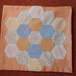 May include: A patchwork cushion featuring a central design of hexagonal shapes in various colours. The hexagons are in shades of white, light blue, and peach, with floral and striped patterns. The cushion has a peach border with a subtle chevron pattern.