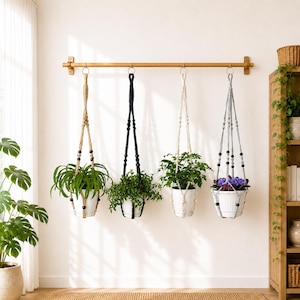 May include: Four macrame plant hangers suspended from a horizontal wooden rod. The hangers feature knotted cord designs in tan, black, off-white, and grey, each holding a white pot containing a green plant, with the last pot holding purple flowers.