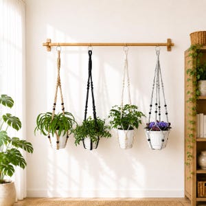 May include: Four macrame plant hangers suspended from a horizontal wooden rod. The hangers feature knotted cord designs in tan, black, off-white, and grey, each holding a white pot containing a green plant, with the last pot holding purple flowers.