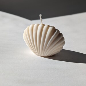 May include: A white seashell-shaped candle with a white wick. The candle is sitting on a white surface.