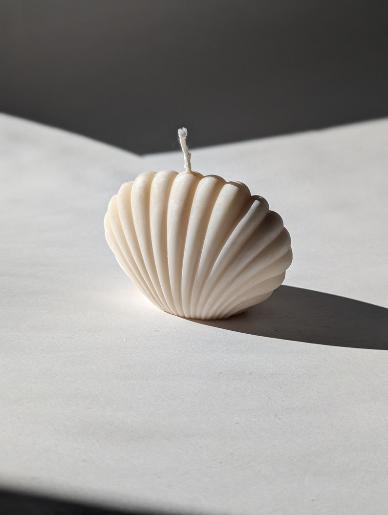 May include: A white seashell-shaped candle with a white wick. The candle is sitting on a white surface.