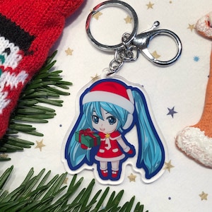 May include: A Christmas-themed keychain featuring a cartoon character with blue hair wearing a red Santa hat and dress, holding a green gift box. The keychain is made of clear acrylic.