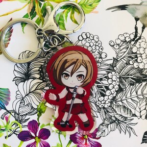 May include: A red and white anime character keychain with a microphone. The character has brown hair and is wearing a red and white outfit. The keychain is attached to a silver key ring.