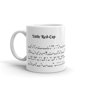 May include: White ceramic mug with a black handle. The mug has the text "Little Red-Cap" printed on it, followed by a line of braille.