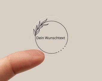 May include: A clear, round sticker with a black line drawing of a circle, a sprig of leaves, and the text "Dein Wunschtext". The sticker is held by a finger against a neutral background.