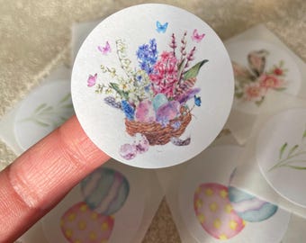 May include: Round sticker featuring a watercolor illustration of an Easter nest with colorful eggs, flowers, and butterflies. The nest is brown, and the eggs are pastel colors. The flowers are pink, blue, and white.