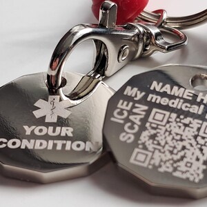 May include: A silver medical alert keychain with two hexagonal tags. One tag has a medical symbol and the words "YOUR CONDITION". The other tag has a QR code and the words "NAME HERE, My medical, ICE SCAN". A red cord is attached.