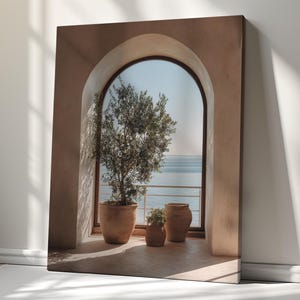 Op de afbeelding: Een rechthoekige canvasprint met een boograam en uitzicht op zee. Een grote olijfboom en twee kleinere planten staan in terracotta potten op een betegelde vloer binnen de boog. De scène is verlicht door warm, natuurlijk zonlicht en schaduwen.