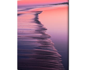 May include: A close-up of a sandy beach at sunset. The sand is wet and rippled, and the water is a soft pink and purple color. The horizon is in the distance, and the sky is a vibrant pink and orange.