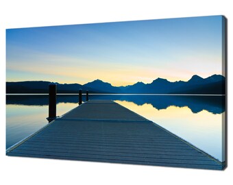 May include: A wooden jetty stretches out into a calm lake, with a mountain range in the distance. The sky is a soft blue with a touch of orange from the setting sun.