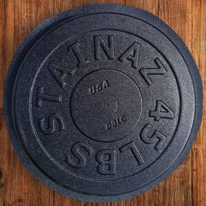 Puede incluir: Una placa de peso de hierro fundido negro con el texto "STAINLESS 45 LBS" y "USA" y "DDLC" impreso en ella.