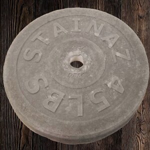 Puede incluir: Una placa de peso de hormig&oacute;n gris con el texto "STAINLESS" y "45 LBS" en relieve en la superficie. La placa tiene un agujero central para una barra.