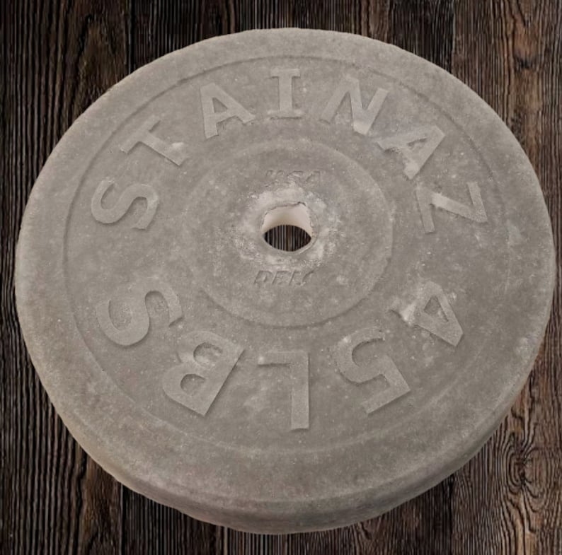 Puede incluir: Una placa de peso de hormig&oacute;n gris con el texto "STAINLESS" y "45 LBS" en relieve en la superficie. La placa tiene un agujero central para una barra.