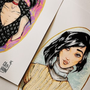 May include: Two watercolor illustrations of women with black hair. The first woman is wearing a black and white patterned top with a red choker. She is surrounded by a pink and gold oval with a spiderweb design. The second woman is wearing a yellow sweater and a white turtleneck. She is surrounded by a blue and gold oval.