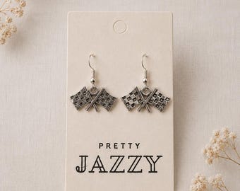May include: A pair of silver-toned dangle earrings featuring a crossed checkered flag design. The earrings are attached to a rectangular cream-colored display card with the text PRETTY JAZZY printed in black lettering on the front surface.