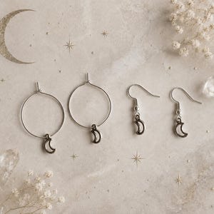 May include: Two silver hoop earrings and two silver hook earrings, each featuring a small crescent moon charm. The jewellery is arranged in a row, showcasing the metallic finish and the simple, circular design of the hoops and the curved hooks.