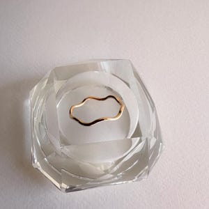 May include: A gold-colored, wavy ring rests inside a clear, faceted, glass container. The ring has a delicate, organic shape. The container is set against a white background, creating a minimalist aesthetic.