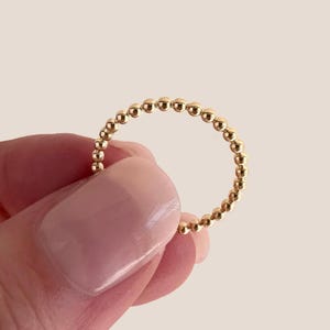 May include: A gold beaded ring. The ring is composed of small, evenly spaced gold beads, forming a continuous circle. The ring is held by a person with a light skin tone, and the background is a soft, neutral color.