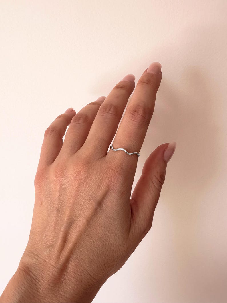 May include: A silver-colored ring with a wavy design. The ring is thin and delicate, with a smooth, polished finish. The ring is worn on a finger, showcasing its simple yet elegant style. The ring's design resembles a small wave.