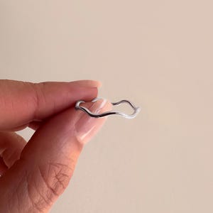 May include: A silver-colored ring with a wavy design. The ring is held by a person. The ring's design is a continuous, flowing line, creating a unique and modern aesthetic. The ring is a piece of jewelry.