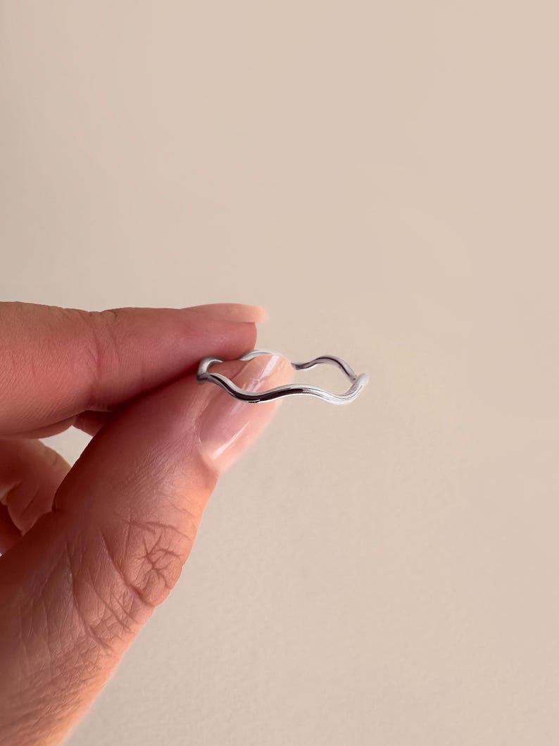 May include: A silver-colored ring with a wavy design. The ring is held by a person. The ring's design is a continuous, flowing line, creating a unique and modern aesthetic. The ring is a piece of jewelry.