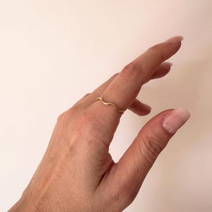 May include: A delicate gold ring with a wavy design. The ring is worn on a finger, showcasing its minimalist and elegant style. The ring's design resembles a gentle wave, adding a touch of sophistication to the jewelry.