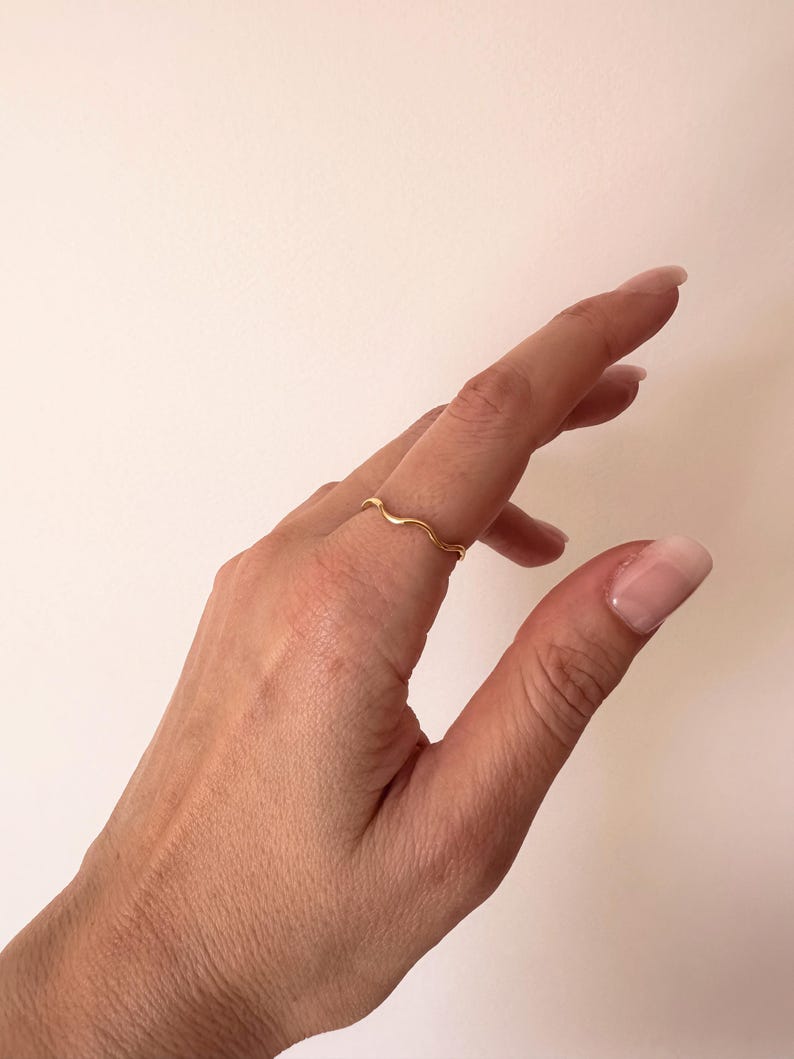 May include: A delicate gold ring with a wavy design. The ring is worn on a finger, showcasing its minimalist and elegant style. The ring's design resembles a gentle wave, adding a touch of sophistication to the jewelry.