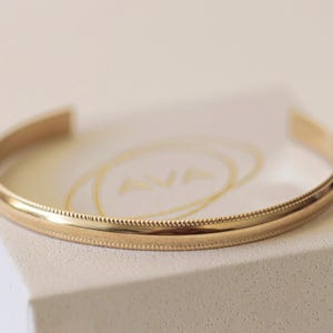 May include: A gold-colored cuff bracelet with a smooth, polished surface and a detailed beaded edge. The open-ended design allows for easy wear. The bracelet is displayed on a white surface.