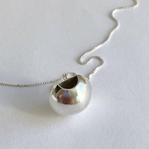 May include: A silver-colored pendant necklace featuring a spherical charm with a hollowed-out top. The charm is attached to a delicate chain. The pendant has a smooth, reflective surface.