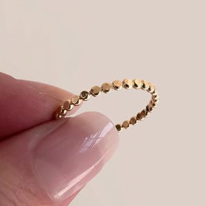 May include: A delicate gold ring featuring a series of small, evenly spaced circular accents. The ring's design is simple and elegant, with a polished finish. The ring is held up against a soft, neutral background.