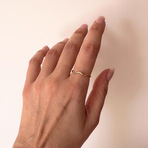 May include: A delicate, gold-colored ring with a wavy design. The ring is worn on a finger, showcasing its minimalist and elegant style. The ring's design resembles a gentle wave.
