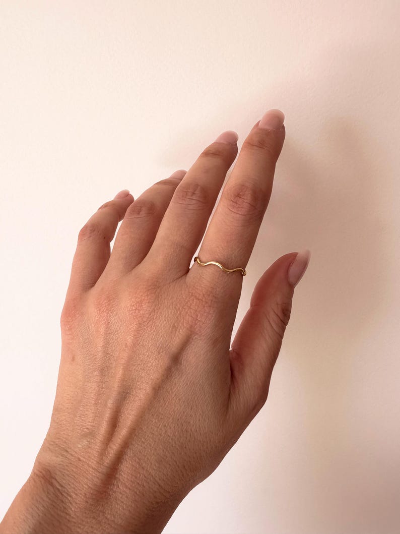 May include: A delicate, gold-colored ring with a wavy design. The ring is worn on a finger, showcasing its minimalist and elegant style. The ring's design resembles a gentle wave.