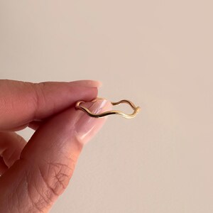 May include: A delicate gold-colored ring with a wavy design. The ring is held between fingers against a neutral background. The ring's design features a continuous, flowing wave pattern.