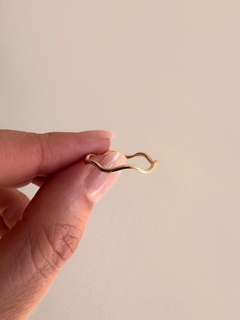 May include: A delicate gold-colored ring with a wavy design. The ring is held between fingers against a neutral background. The ring's design features a continuous, flowing wave pattern.
