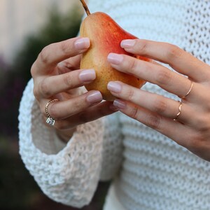 May include: A ripe pear with a red and yellow gradient is held by a person. The pear's stem is brown. The person is wearing rings and a white, open-knit sweater. The pear is the focal point of the image.